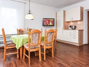 Un comedor con mesa y sillas de madera, televisor en la pared y zona de cocina con armarios y electrodomésticos.