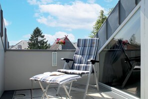 A balcony with a striped chair and a small table, a window, and a view of a house and trees.
