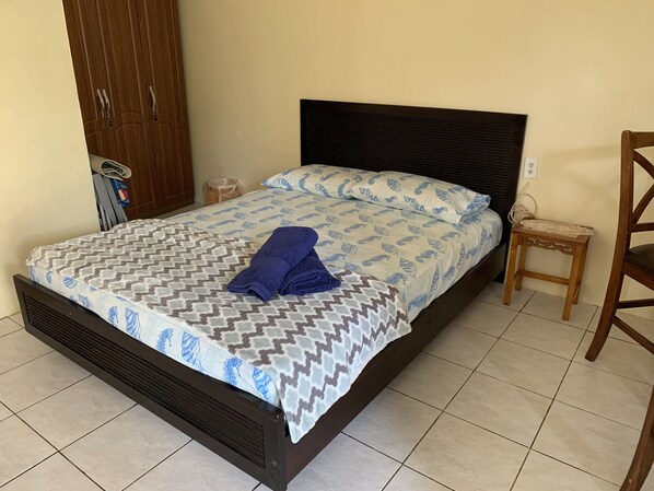 Uma cama com roupa de cama estampada e uma toalha dobrada, uma mesa de cabeceira de madeira com um objeto decorativo, e um guarda-roupa de madeira num quarto com soalho em azulejos.