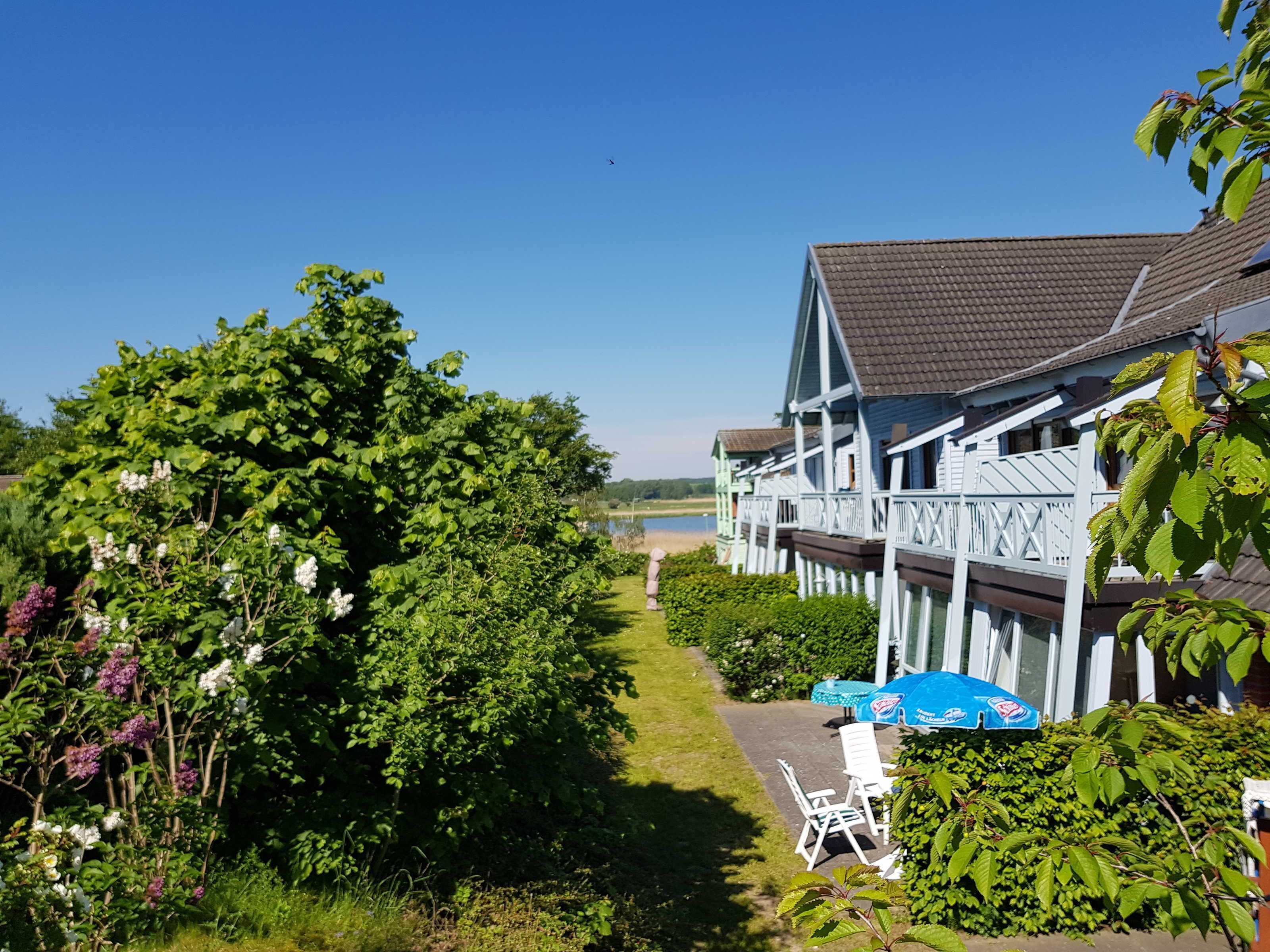 A row of white buildings with a covered walkway, a grassy path, and a body of water in the background.