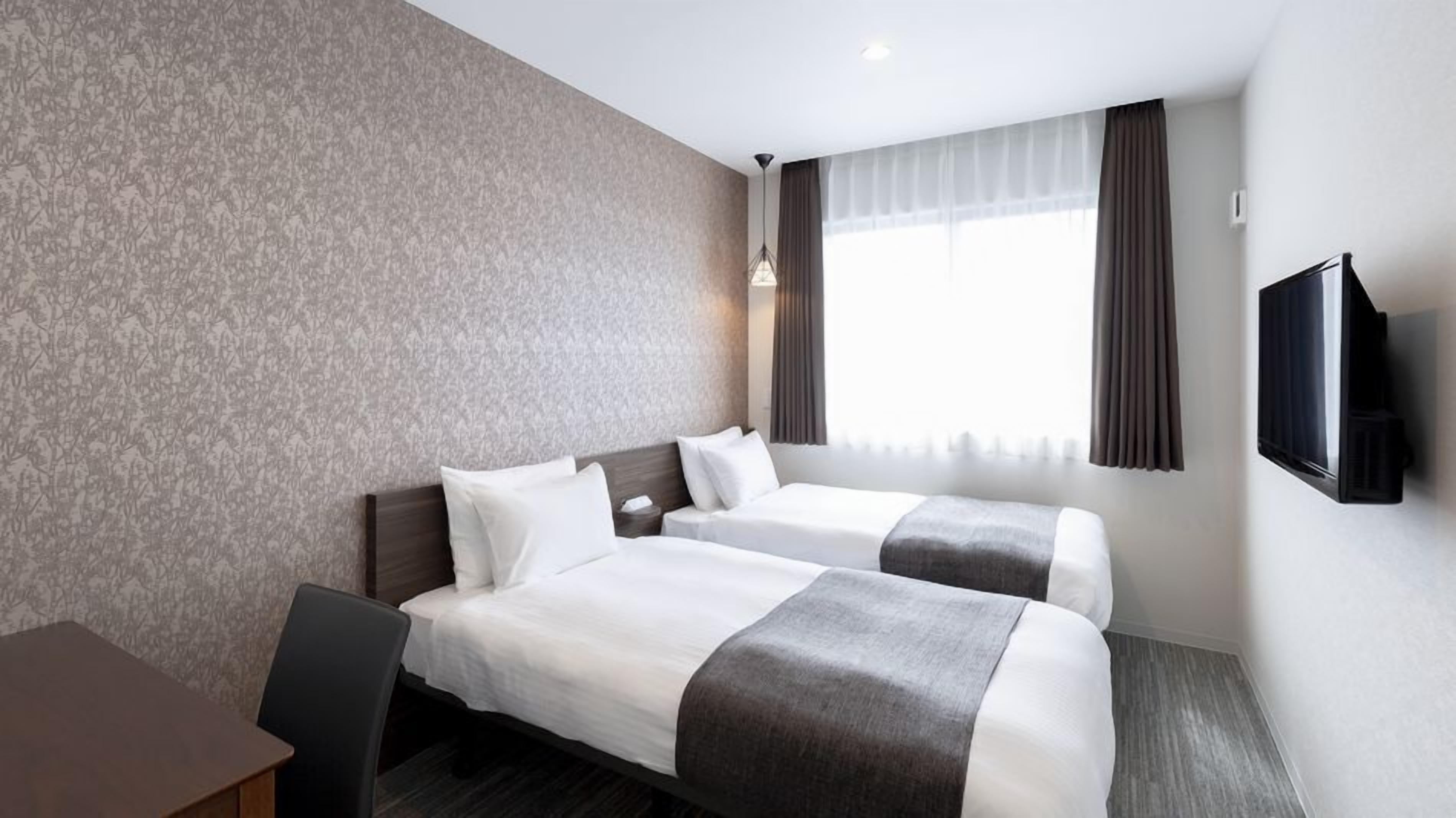 A hotel room with two beds, a desk, a TV, and a window with curtains.