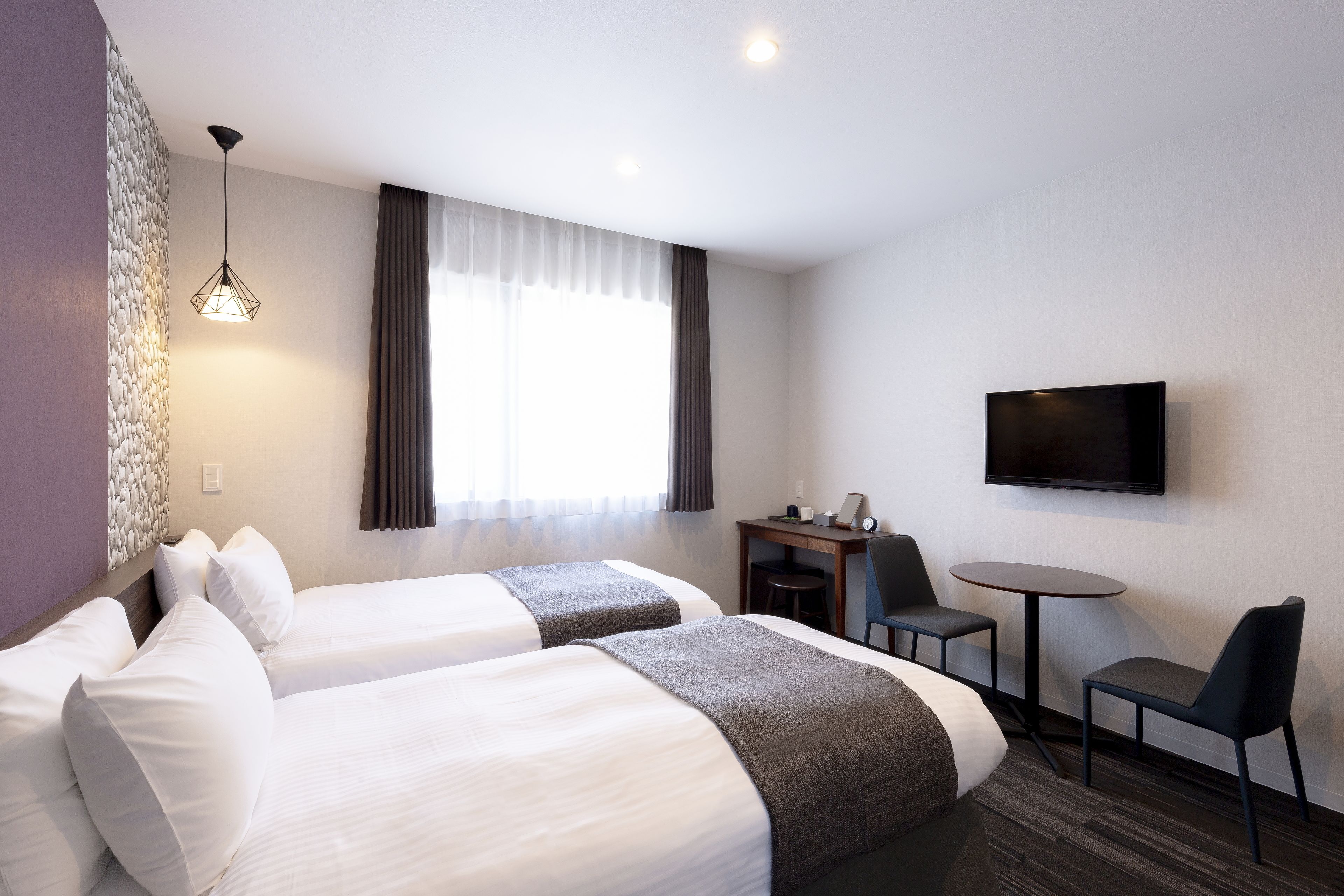 A hotel room with two beds, a desk with a chair, a small table, and a TV mounted on the wall.