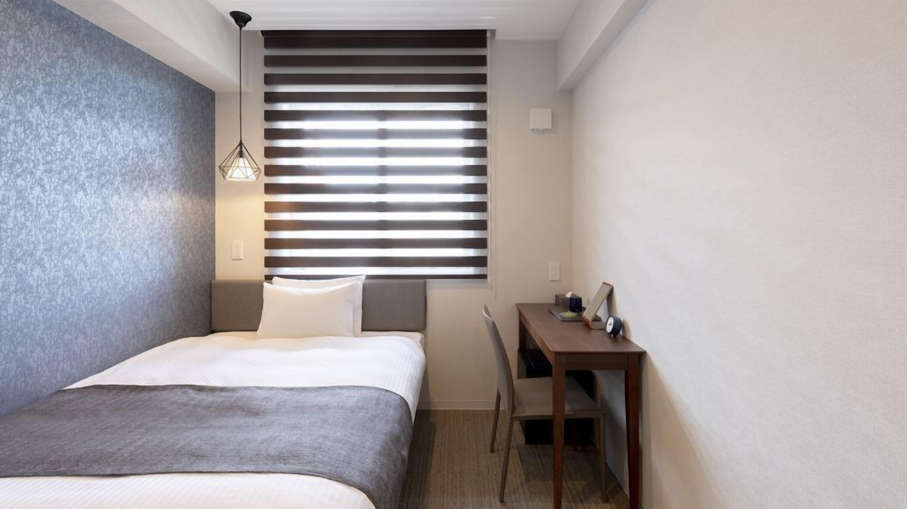 A compact hotel room with a bed, a desk, and a window with blinds.