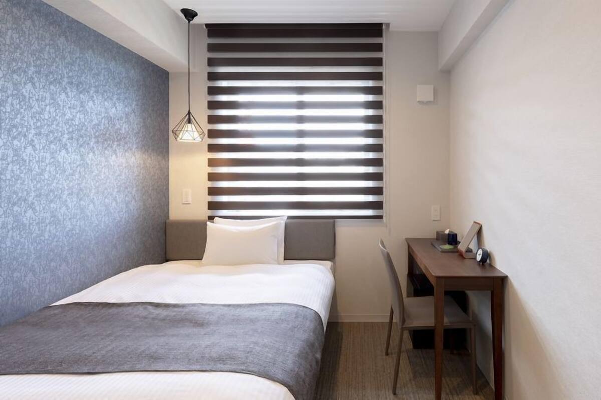 Ein kleines Hotelzimmer mit Bett, Schreibtisch und einem Fenster mit Jalousien.