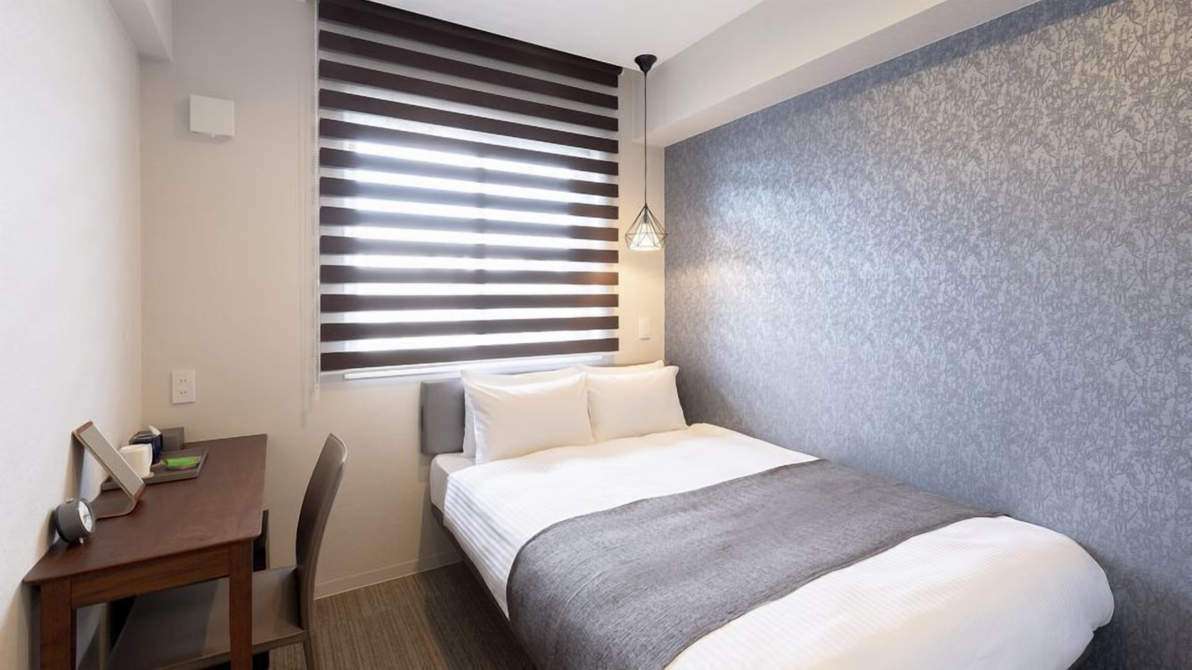 A hotel room with a bed, a desk, and a window with blinds.