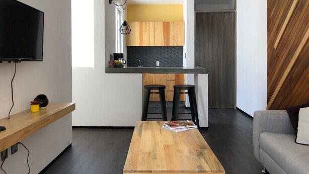 Una cuina moderna amb armaris de fusta, un taulell negre i forn integrat. Hi ha una zona de menjador amb taula de fusta i dos tamborets negres. Un televisió de pantalla plana està penjat a la paret.