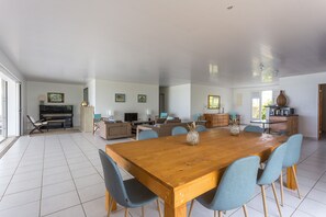 A spacious living area with a large wooden dining table surrounded by chairs, a sofa set, and a television.