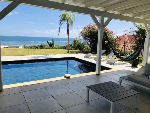 A pool area with a view of the ocean, a hammock, and a patio with a bench.
