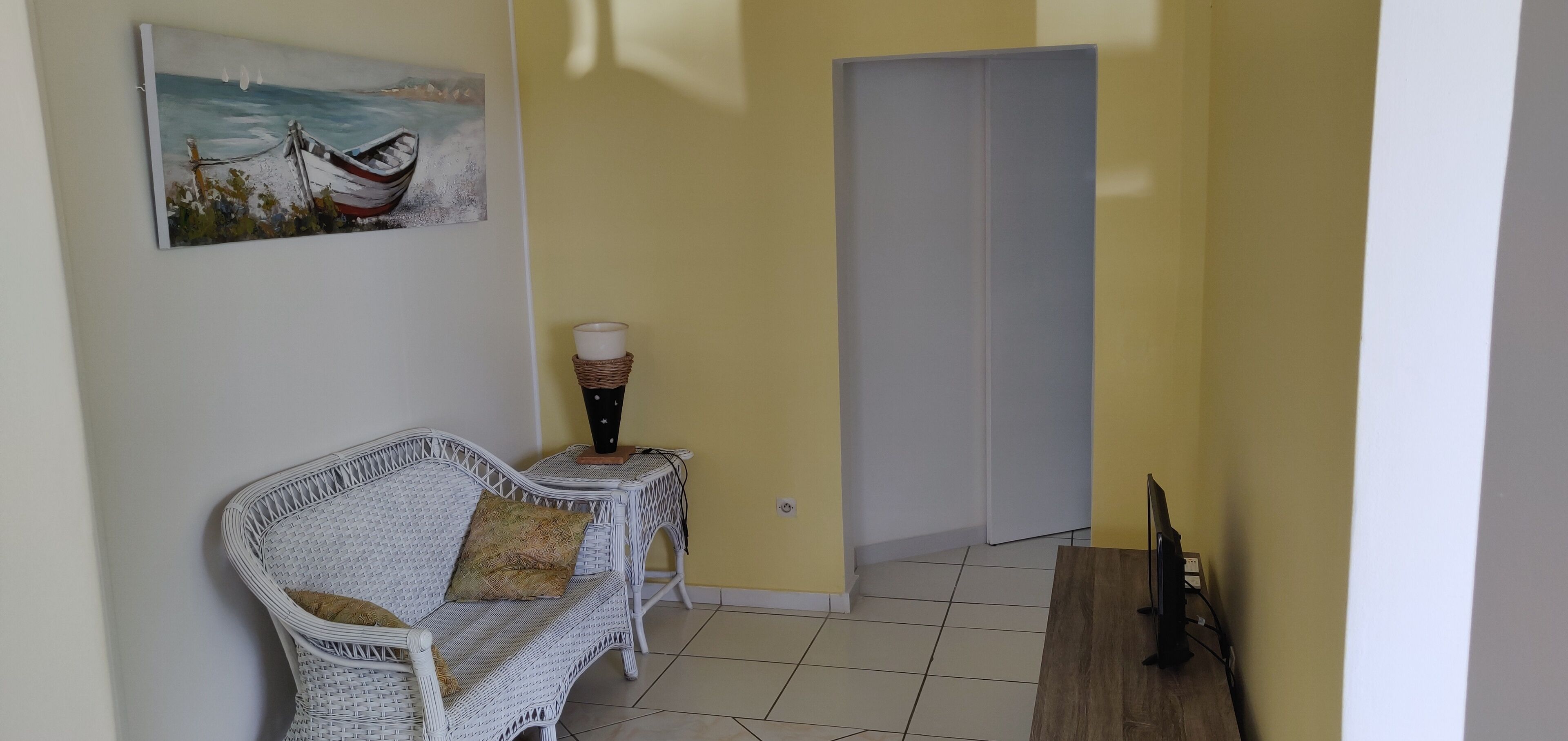 Une pièce avec un canapé en osier blanc, une petite table d’appoint sur laquelle est posée une bougie, et un tableau représentant un bateau accroché au mur.
