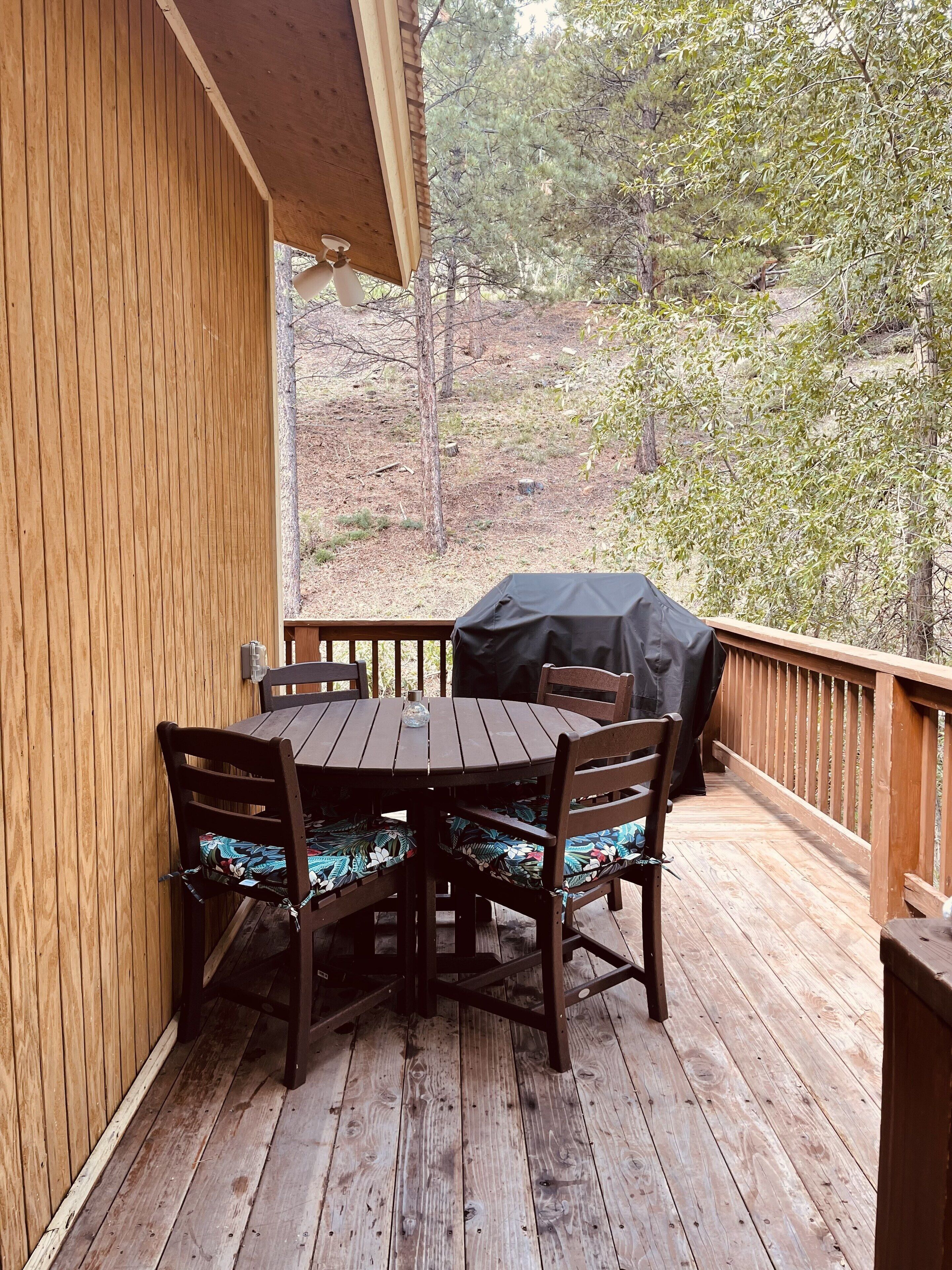 A wooden deck with a round table and chairs, a barbecue grill, and a view of a wooded area.