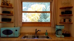 A kitchen with a window, a sink, a retro-style microwave, a coffee maker, and shelves with cups and mugs.