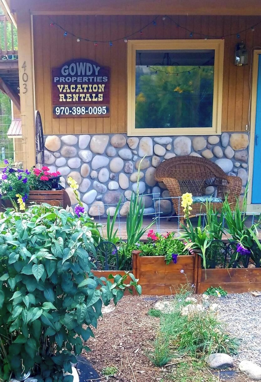 Une maison de vacances avec un mur en pierre, une chaise en osier et une enseigne indiquant « Gowdy Properties ».