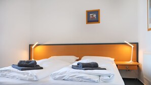 Un letto matrimoniale rifatto con cura, lenzuola bianche e due asciugamani piegati, una testiera in legno, un comodino con una lampada e un quadro incorniciato appeso alla parete.