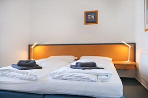 A neatly made double bed with white linens and two folded towels, a wooden headboard, a bedside table with a lamp, and a framed picture on the wall.