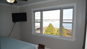 A bedroom with a large window, a bed, a TV mounted on the wall, and a view of a body of water and trees.