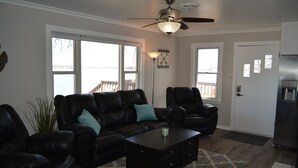 A living room with a black leather sofa, a matching armchair, a coffee table, and a ceiling fan. There are large windows providing a view of the outdoors.