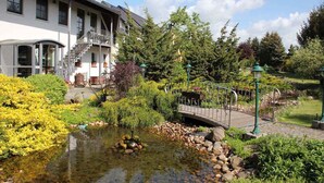 A building with a staircase leading to a balcony, surrounded by lush greenery and a small pond with a bridge.