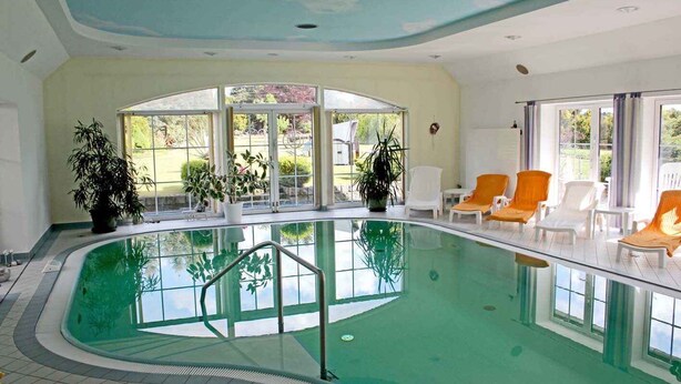 An indoor swimming pool with clear glass walls, surrounded by lounge chairs and potted plants.