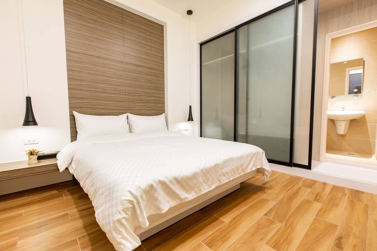 Un dormitorio moderno con una cama grande, cabecera de madera, puerta de vidrio corredera y baño integrado con lavamanos y espejo.
