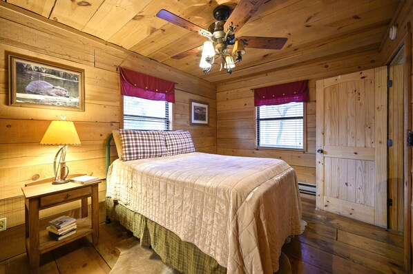 Ein Schlafzimmer in einer Holzhütte mit einem Bett, einem Nachttisch mit Lampe, einem Deckenventilator und einem Fenster mit Vorhang.