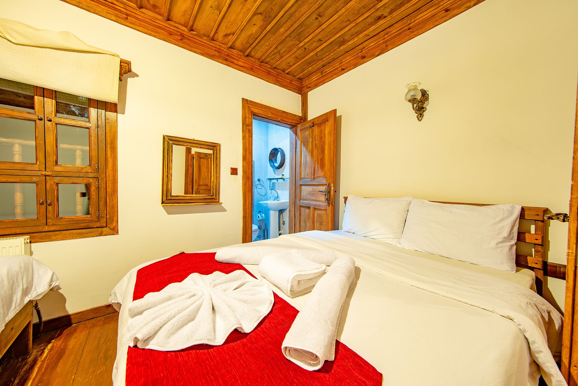 A hotel room with a wooden ceiling, a bed with white linens, a red throw blanket, and a wooden wardrobe.