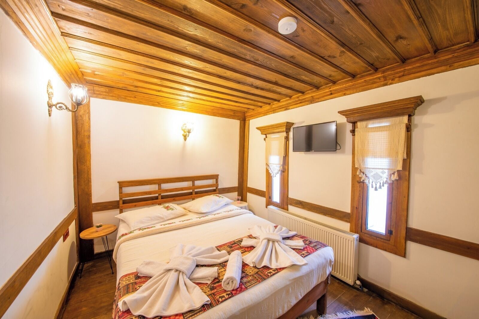 A bedroom with a wooden ceiling, a bed with a patterned bedspread, two wall-mounted lights, a television, and a window with a curtain.