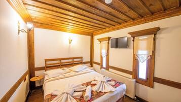 A bedroom with a wooden ceiling, a bed with a patterned bedspread, two wall-mounted lights, a television, and a window with a curtain.