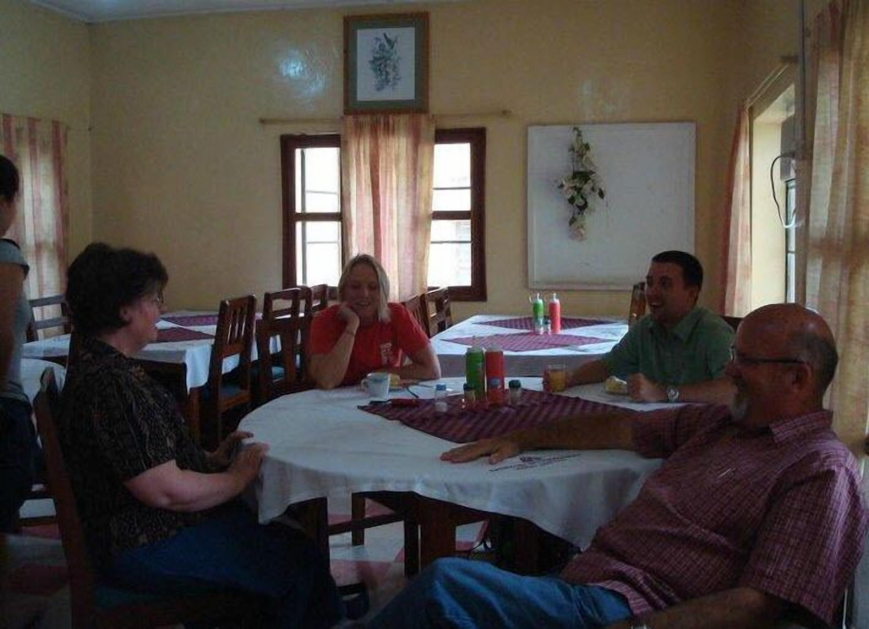 Um grupo de pessoas sentadas à volta de uma mesa numa sala com decoração simples, destacando-se uma janela com cortinas, um ornamento fixado na parede e uma mesa com suporte para prato.