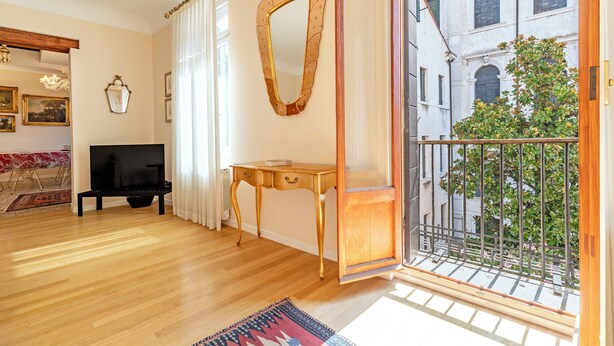 A living room with wooden flooring, a TV stand, a table, a mirror, and a balcony with a view of a building and greenery.