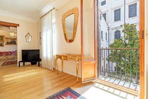 Een woonkamer met houten vloeren, een tv-meubel, een tafel, een spiegel, en een balkon met uitzicht op een gebouw en groen.