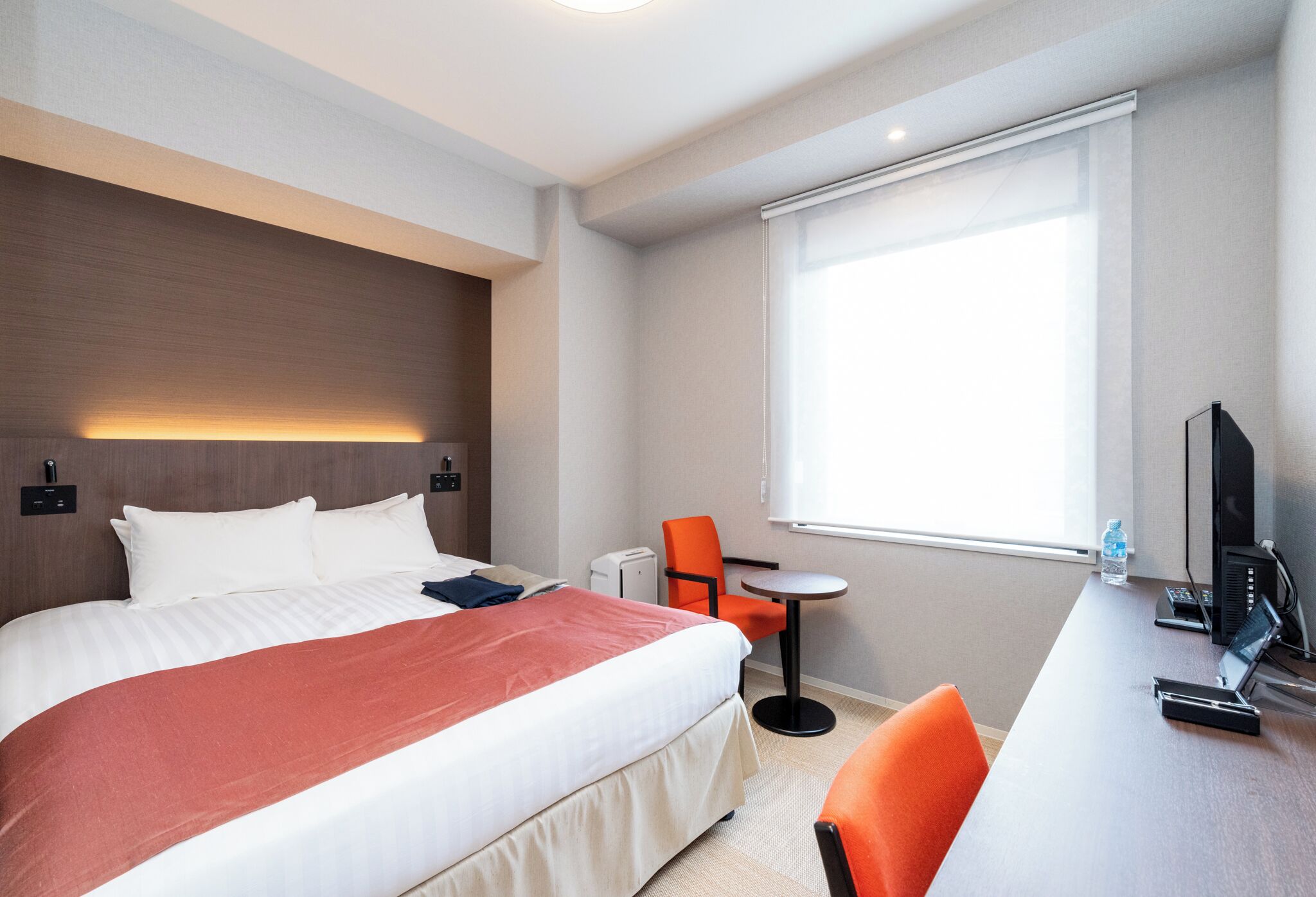 Ett hotellrum med en stor säng, ett skrivbord med en dator, en stol och ett fönster med persienner.