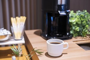 A coffee maker, a cup of coffee, and a bowl of straws on a wooden table.