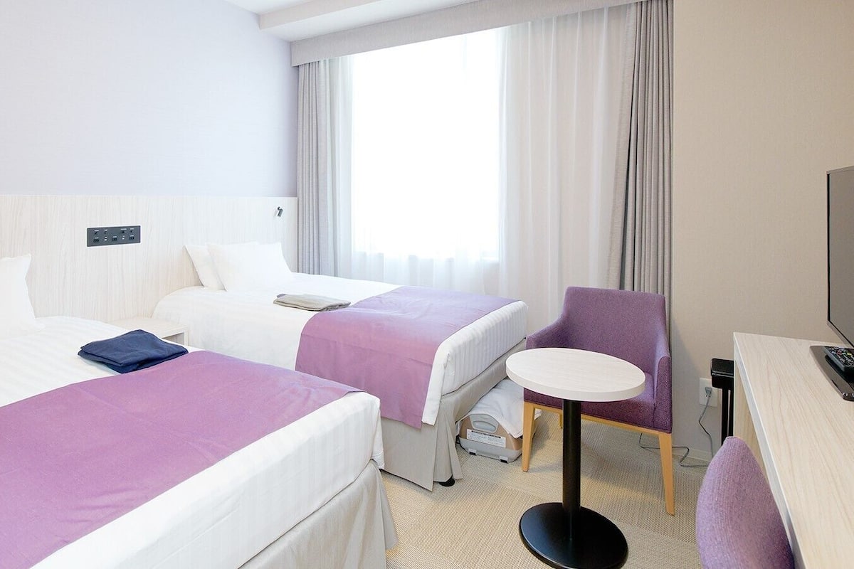 Ein Hotelzimmer mit zwei Betten, einem Schreibtisch, einem Stuhl, einem Fernseher und einem Fenster mit Vorhängen.