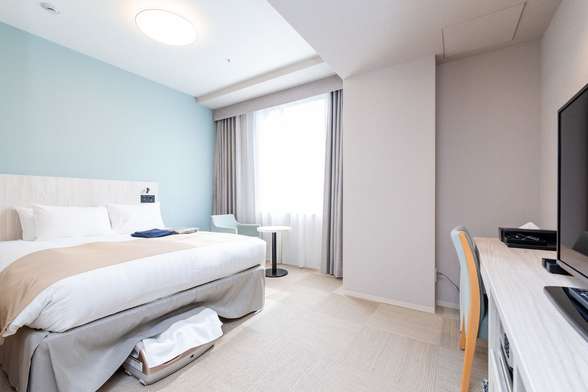 Ein Hotelzimmer mit Bett, Schreibtisch mit Computer, Fernseher, Fenster mit Vorhängen und einem kleinen Tisch.
