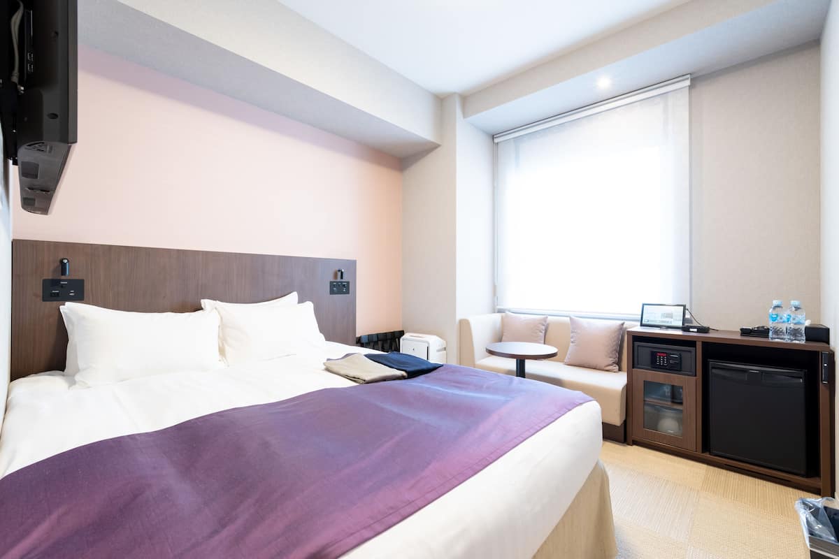 Ein Hotelzimmer mit Bett, Schreibtisch mit Computer, einer Minikühlschrank, einer Kaffeemaschine und einem Fenster mit Jalousien.