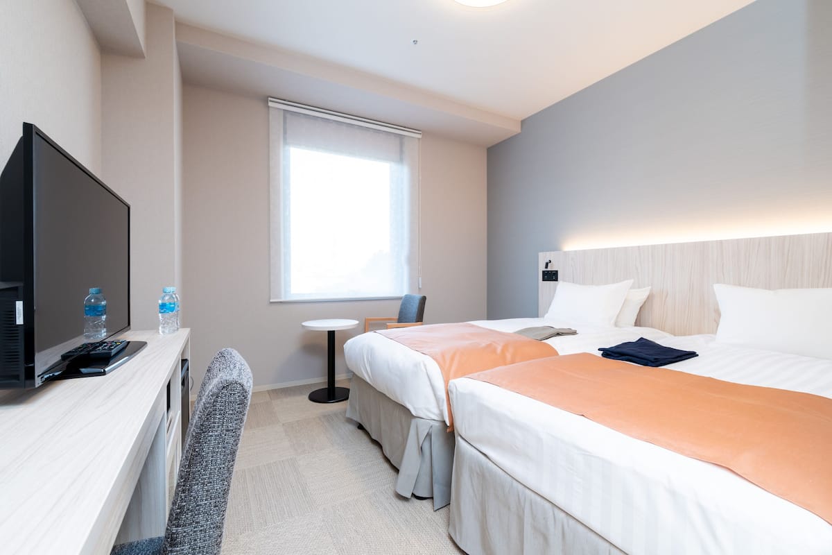 Ein Hotelzimmer mit zwei Betten, einem Schreibtisch mit Fernseher und Wasserflaschen, einem kleinen Tisch und einem Fenster mit Jalousien.