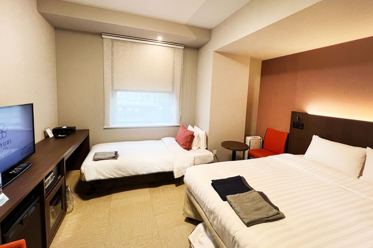 Ein Hotelzimmer mit zwei Betten, einem Schreibtisch mit Fernseher, einem kleinen Tisch und einem Fenster mit Jalousien.