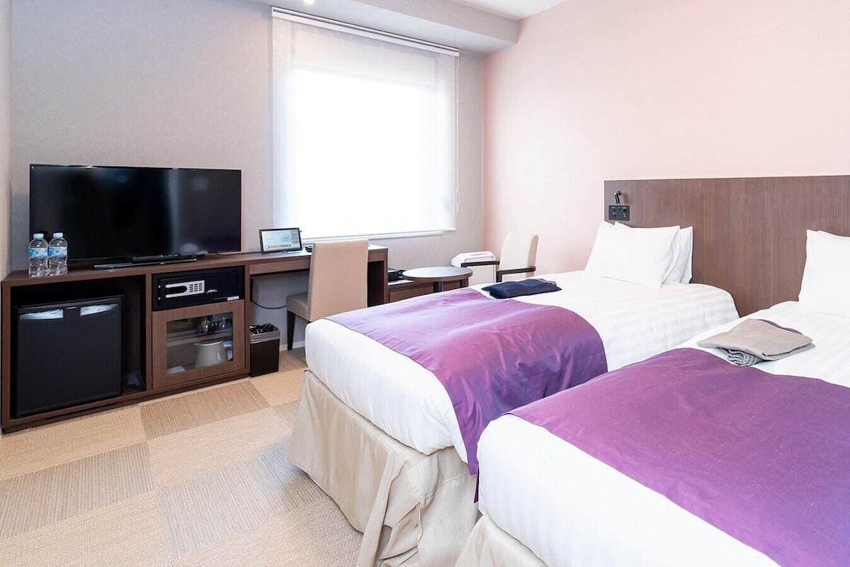 Ein Hotelzimmer mit zwei Betten, einem Fernseher, einem Schreibtisch und einer kleinen Küchenzeile.