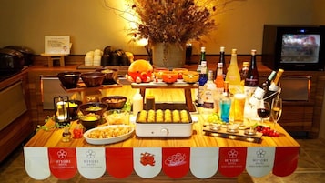 Mesa de buffet con diferentes alimentos y bebidas, un televisor al fondo y un cartel que indica el nombre del hotel.
