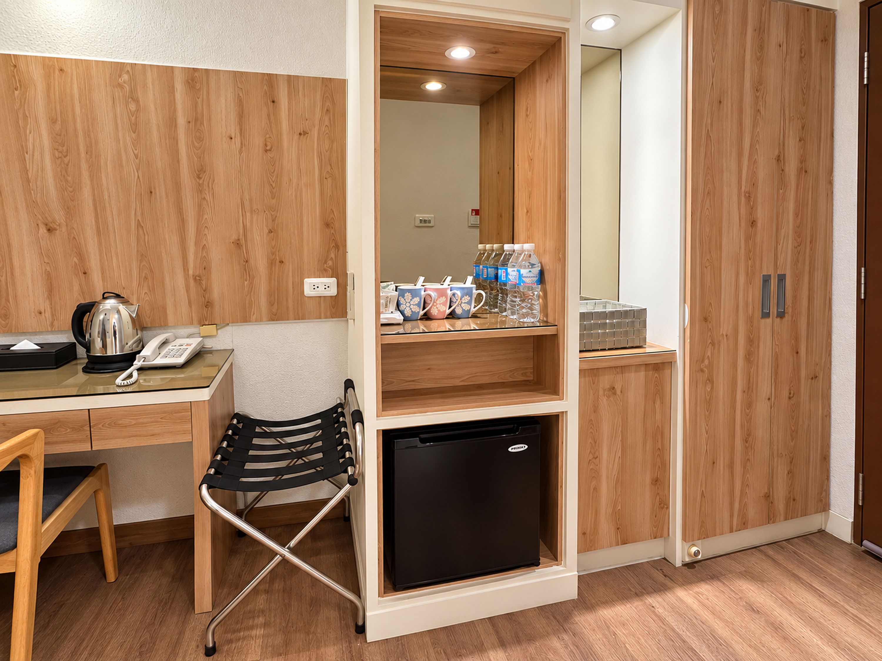 A compact room with a wooden desk, a black chair, a small black refrigerator, and a shelf with bottled water and cups.