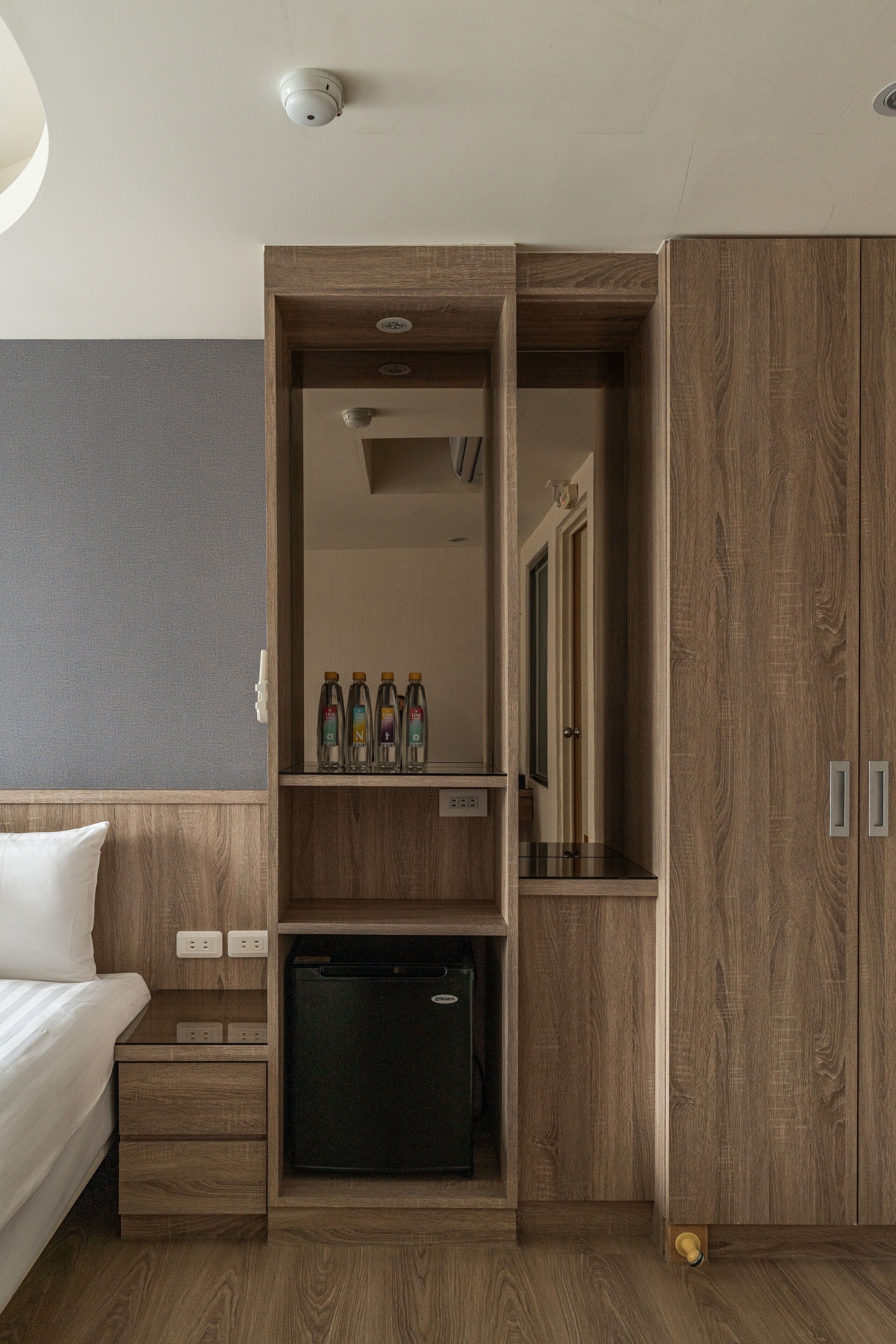 A hotel room with a wooden wardrobe, a small refrigerator, a bedside table with a lamp, and a bed.