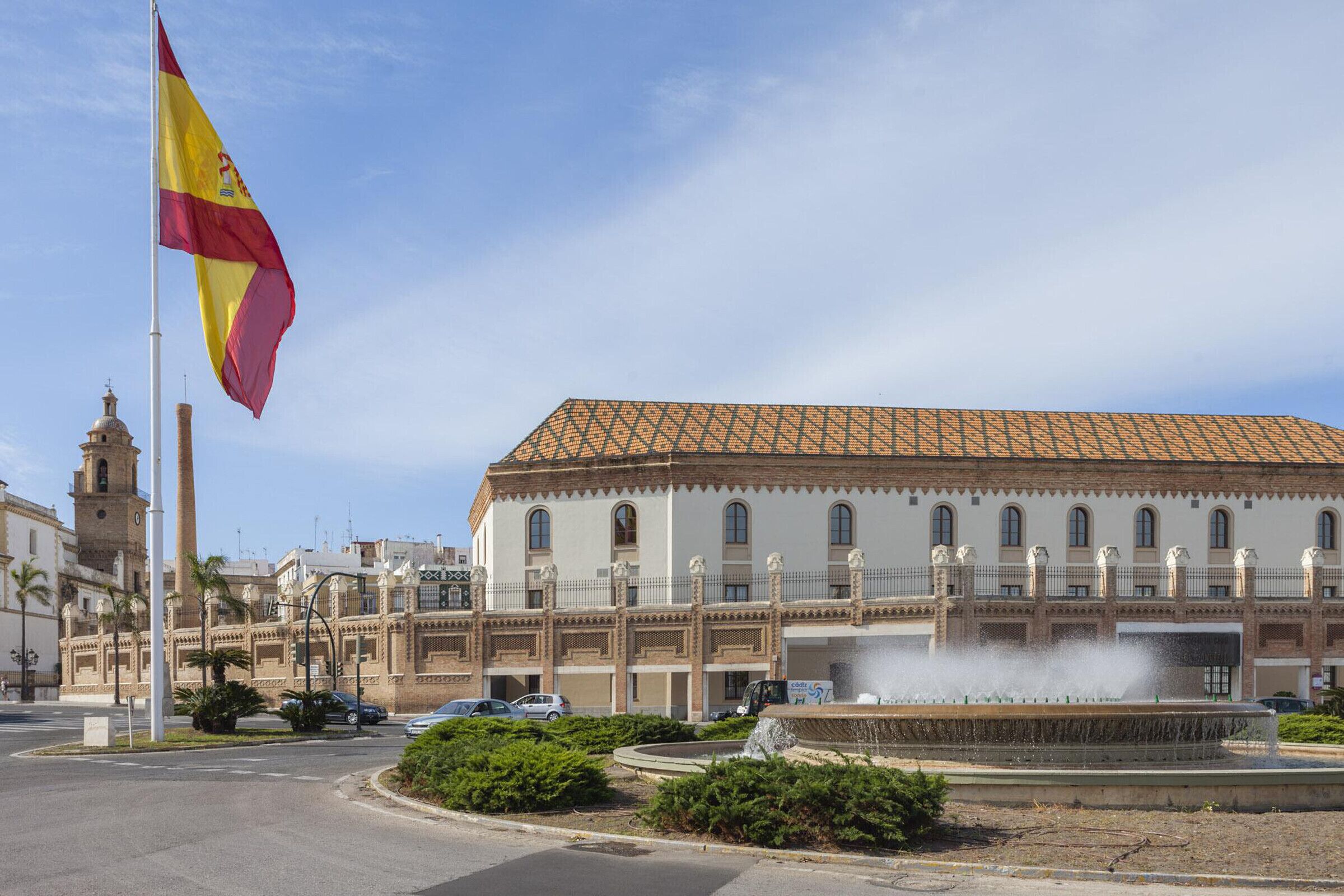 A historic building with a tiled roof, a fountain in the foreground, and a flagpole with a flag.