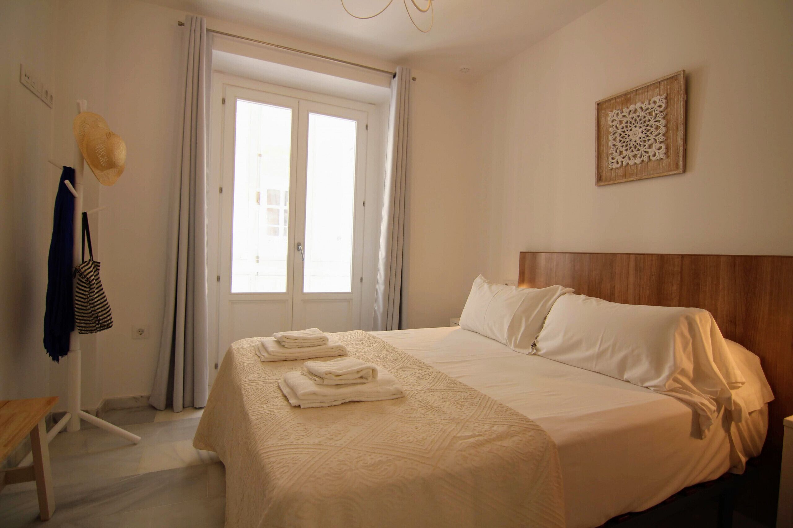 Une chambre d’hôtel avec un lit, une tête de lit en bois, une porte avec des rideaux, un élément décoratif fixé au mur et un banc sur lequel est posée une serviette.