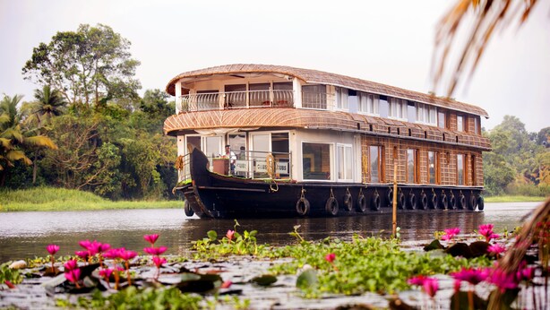 Ein Hausboot mit Holldeck und mehreren Fenstern, das auf einem ruhigen Fluss zwischen üppigem Grün und rosa Seerosen treibt.