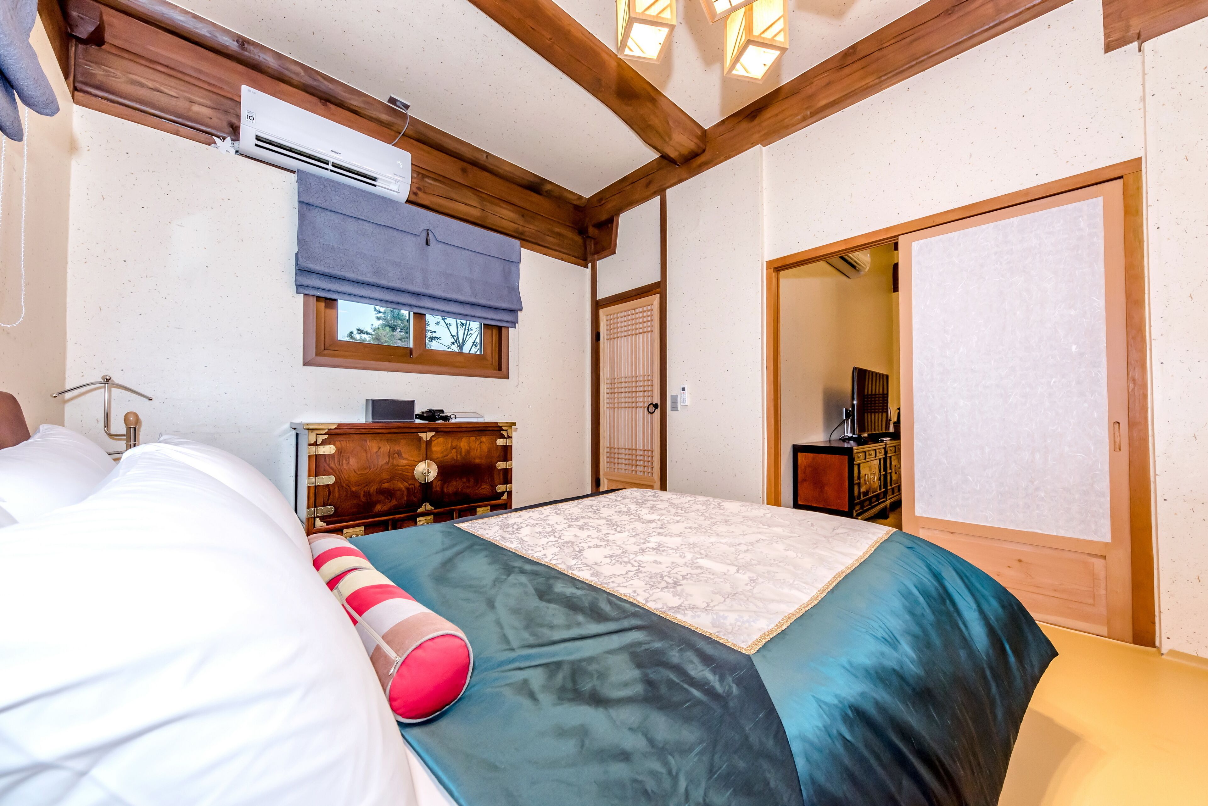 A bedroom with a bed, a wooden dresser, an air conditioning unit, a window with a blue blind, and a door leading to another room.