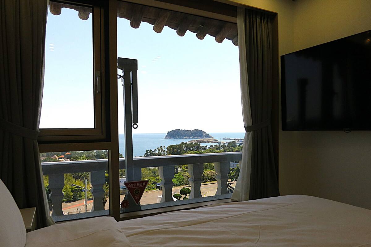Habitación de hotel con una cama, un televisor montado en la pared y un balcón con vistas al mar y una isla distante.