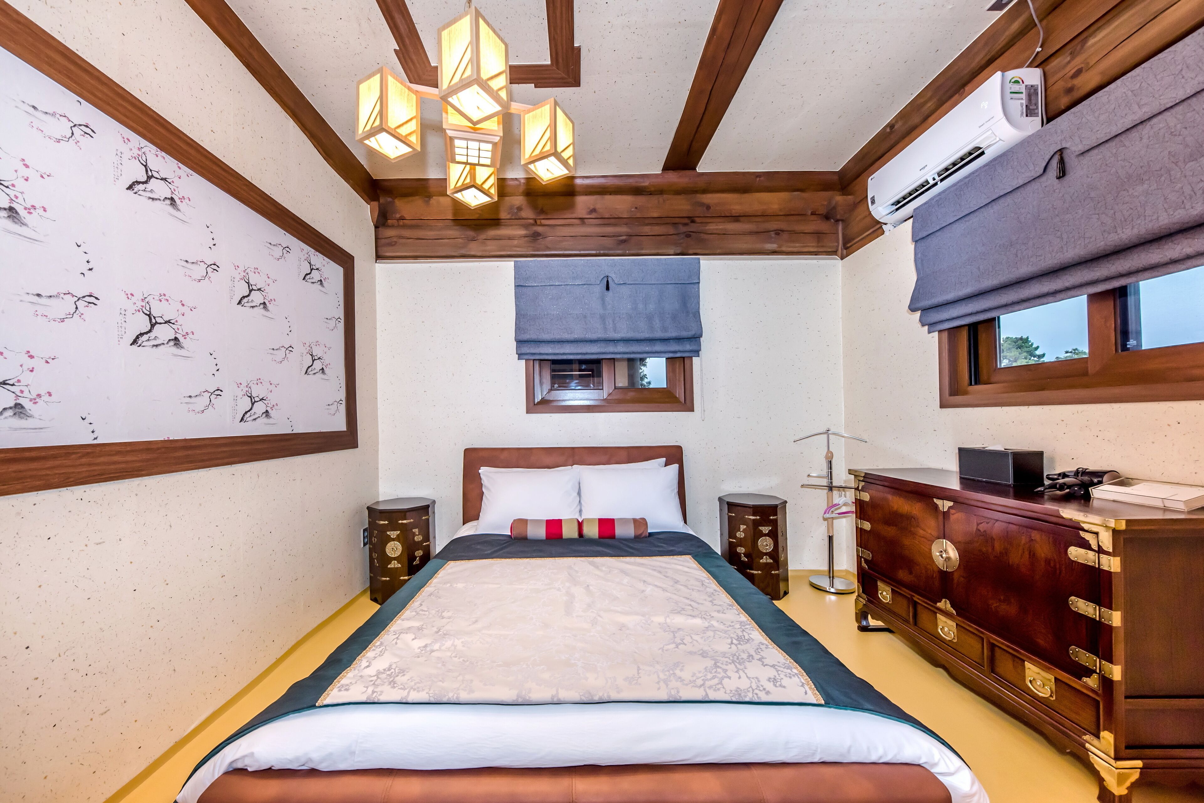 A bedroom with a large bed, wooden ceiling beams, a wall-mounted air conditioner, a bedside table with a lamp, and a wooden dresser.