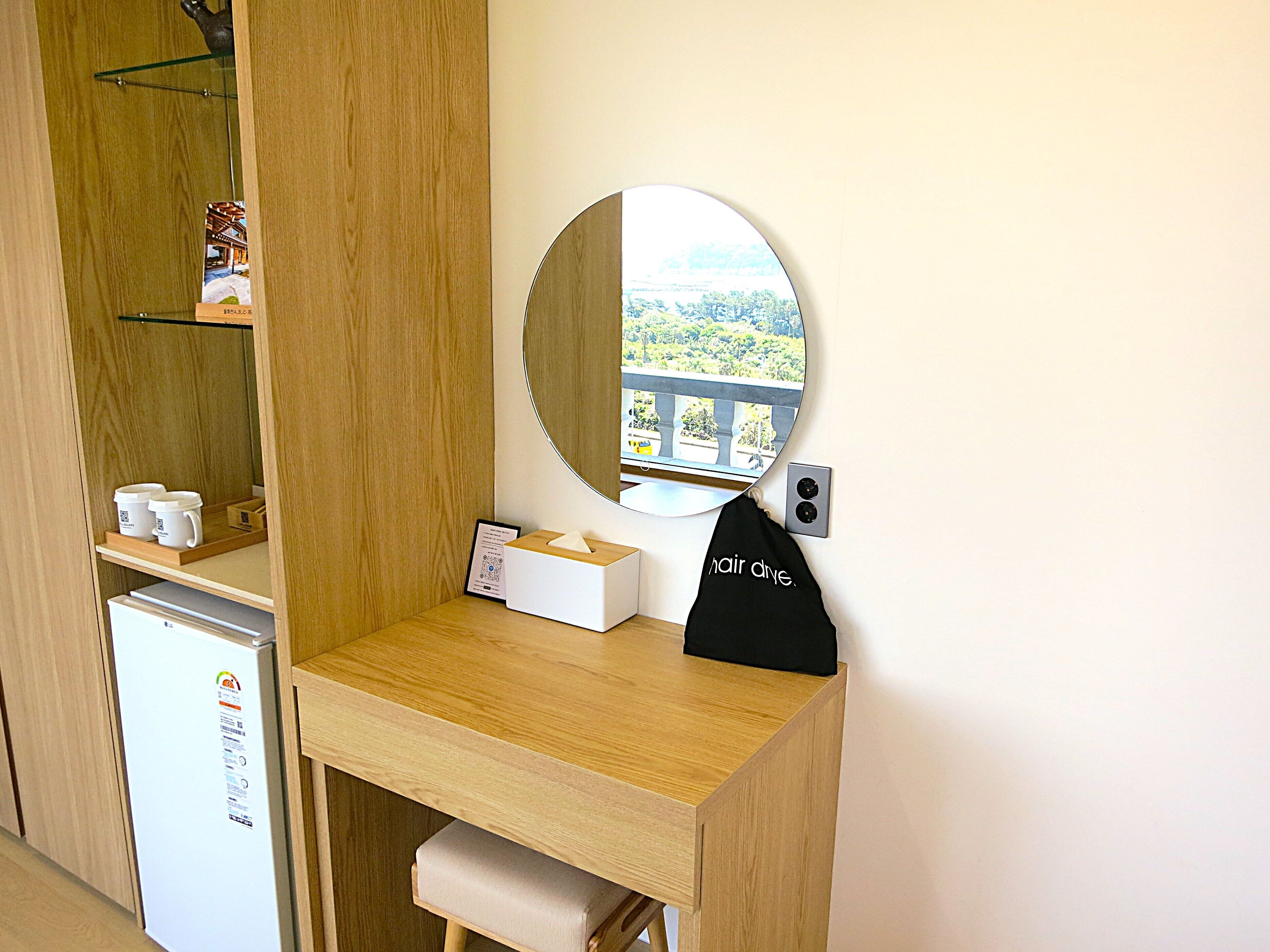 A compact room with a wooden desk, a small refrigerator, a mirror, and a view of greenery.