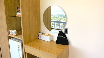 A compact room with a wooden desk, a small refrigerator, a mirror, and a view of greenery.