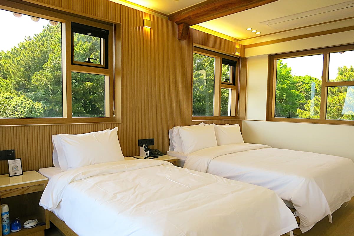 Habitación de hotel con dos camas, paneles de madera y ventanas amplias con vista a follaje.